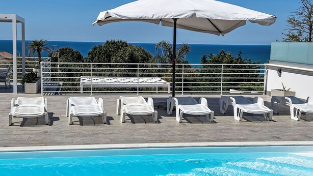 Ein Poolbereich mit Liegestühlen, einem weißen Sonnenschirm und Blick auf den Ozean.