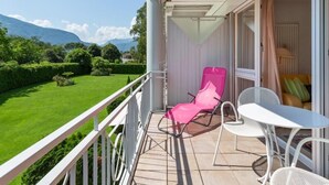 Un balcon donnant sur une pelouse verdoyante et des montagnes, avec une chaise rose, une table blanche et un canapé jaune.