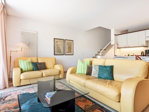 A living room with two beige sofas, a glass coffee table, and a patterned rug. A staircase leads to a kitchen area in the background. - Golf Ranch, 1er Stock by Interhome (Ascona)