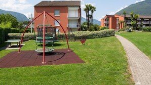 Une aire de jeux avec des balançoires et un toboggan, entourée d’une pelouse bien entretenue et d’un chemin pavé.