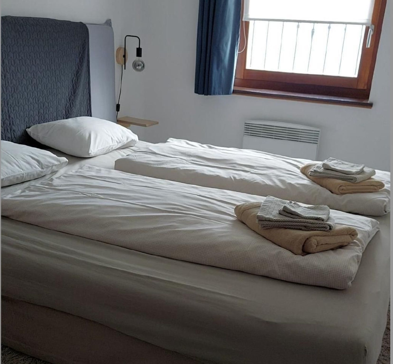 Habitación de hotel con dos camas individuales, una mesita de noche con lámpara, una ventana con cortinas y un calentador de pared.