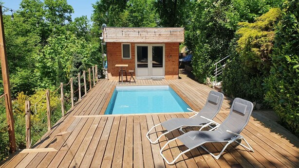 En træterrasse med swimmingpool, to liggestole og en mindre træbygning med dør og vinduer.