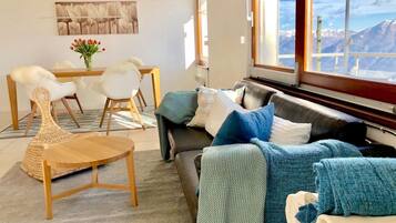Ein modernes Wohnzimmer mit Sofa, Sessel, einem kleinen Tisch und Blick auf Berge durch große Fenster.
