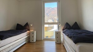 A bedroom with two beds, a window with a mountain view, and a small table. - Wohnung Orizia by Interhome (Locarno)
