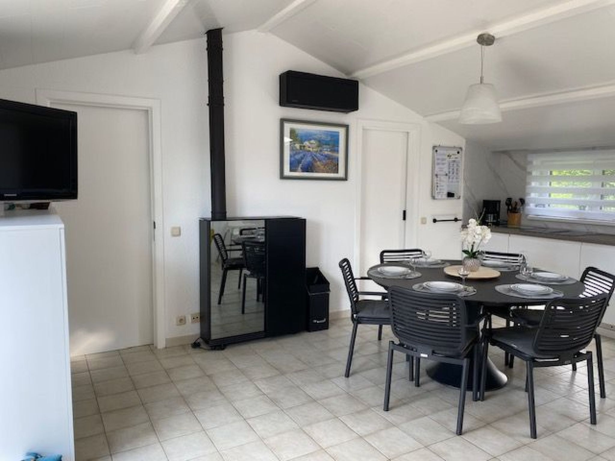 A dining area with a black table set for four, surrounded by black chairs. A large mirror hangs on the wall, and a television is mounted above the table.