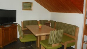 Un área de comedor con una mesa de madera y sillas tapizadas de verde, un televisor sobre un mueble de madera, y un cuadro en la pared.