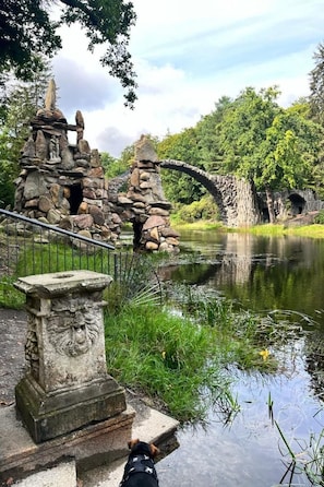 Eine steinerne Brücke über einen stillen Teich, umgeben von üppigem Grün und einem Steinbauwerk mit einer Statue.