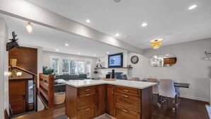 A modern kitchen with wooden cabinets, a marble countertop, and a dining area with white chairs. - The Stag House : HotTub, Outdoor Theatre, Arcade Games, Big Backyard & Sleeps 18 (Langley)
