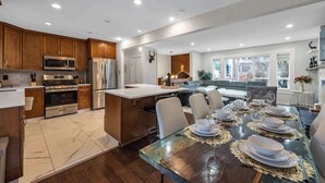 A modern kitchen with wooden cabinets, stainless steel appliances, and a dining area with a glass table set for six. - The Stag House : HotTub, Outdoor Theatre, Arcade Games, Big Backyard & Sleeps 18 (Langley)
