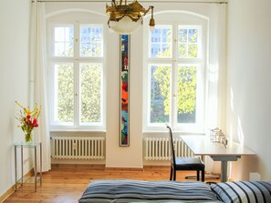 A room with large windows, a radiator, a table, a chair, a bed, and a vase with flowers. - Altbauwohnung in Berlin by Interhome (Berlin)