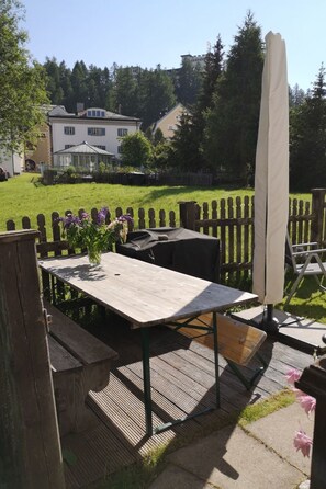 Eine Terrasss mit Holztisch und -stühlen, einem weißen Sonnenschirm und Blick auf ein Haus und Bäume im Hintergrund.