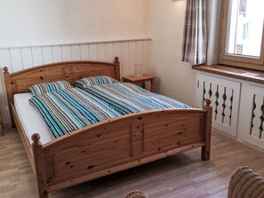 Ein hölzernes Bett mit gestreifter Bettdecke, ein Nachttisch, ein Fenster mit Vorhängen und eine Heizung.