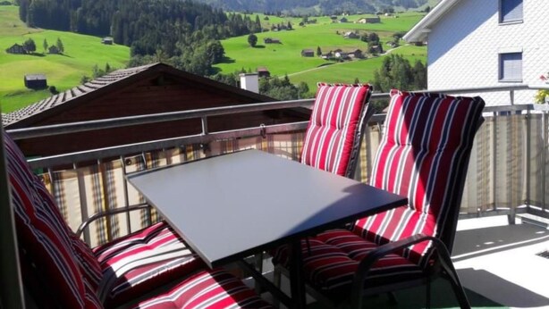 Ein Balkon mit Tisch und gestreiften Stühlen, der auf eine grüne Landschaft mit Gebäuden und Bergen blickt.