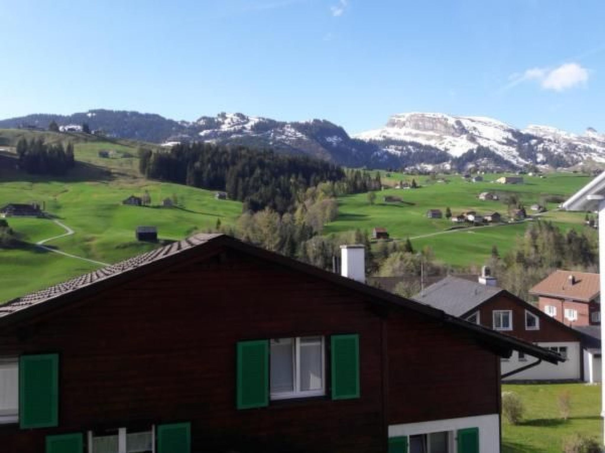 Una casa de madera con contraventanas verdes, rodeada de campos verdes y montañas al fondo.