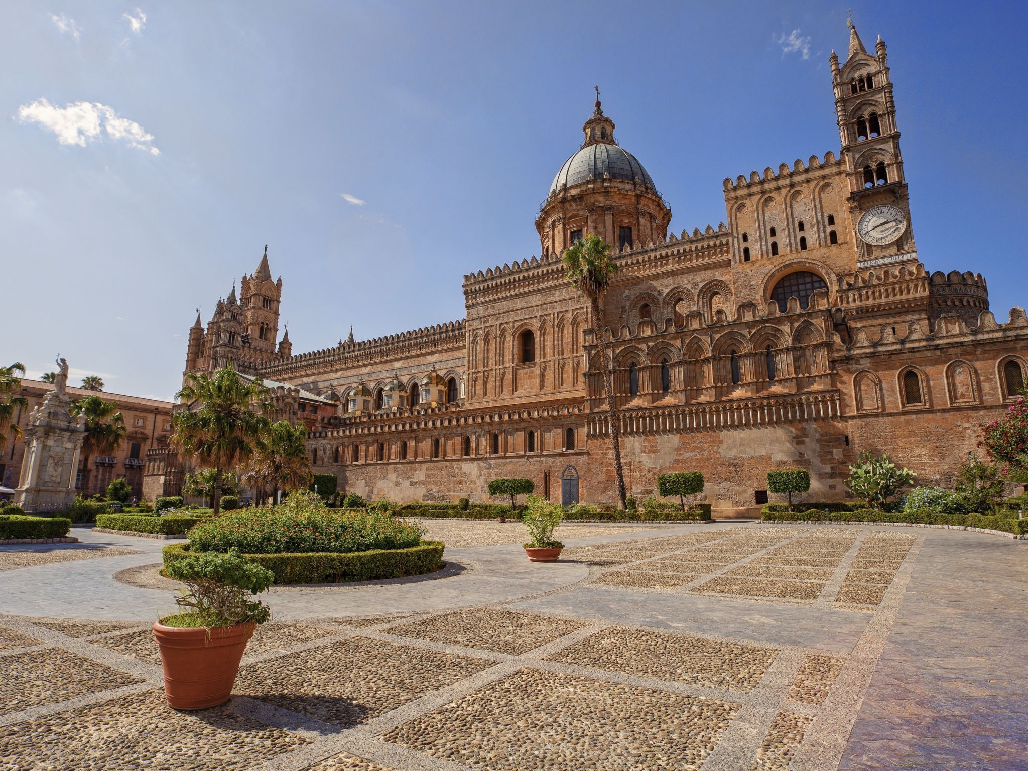 A historic building with a large dome and clock tower, surrounded by a courtyard with potted plants and stone paving.
