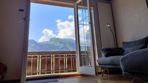 A room with a sliding glass door leading to a balcony, a grey sofa, and a wooden ceiling. - im Gründli by Interhome (Wildhaus)