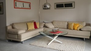 Een woonkamer met een beige bank, een glazen salontafel, een vloerlamp en een vloerkleed.