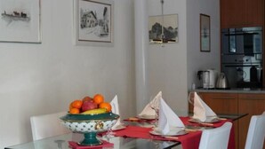 Een eethoek met een glazen tafel, witte stoelen, een schaal fruit en ingelijste foto's aan de muur.