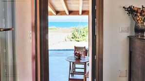 Una puerta que da a un patio con mesa y sillas, vista a la playa y un jarrón con flores sobre un mueble.