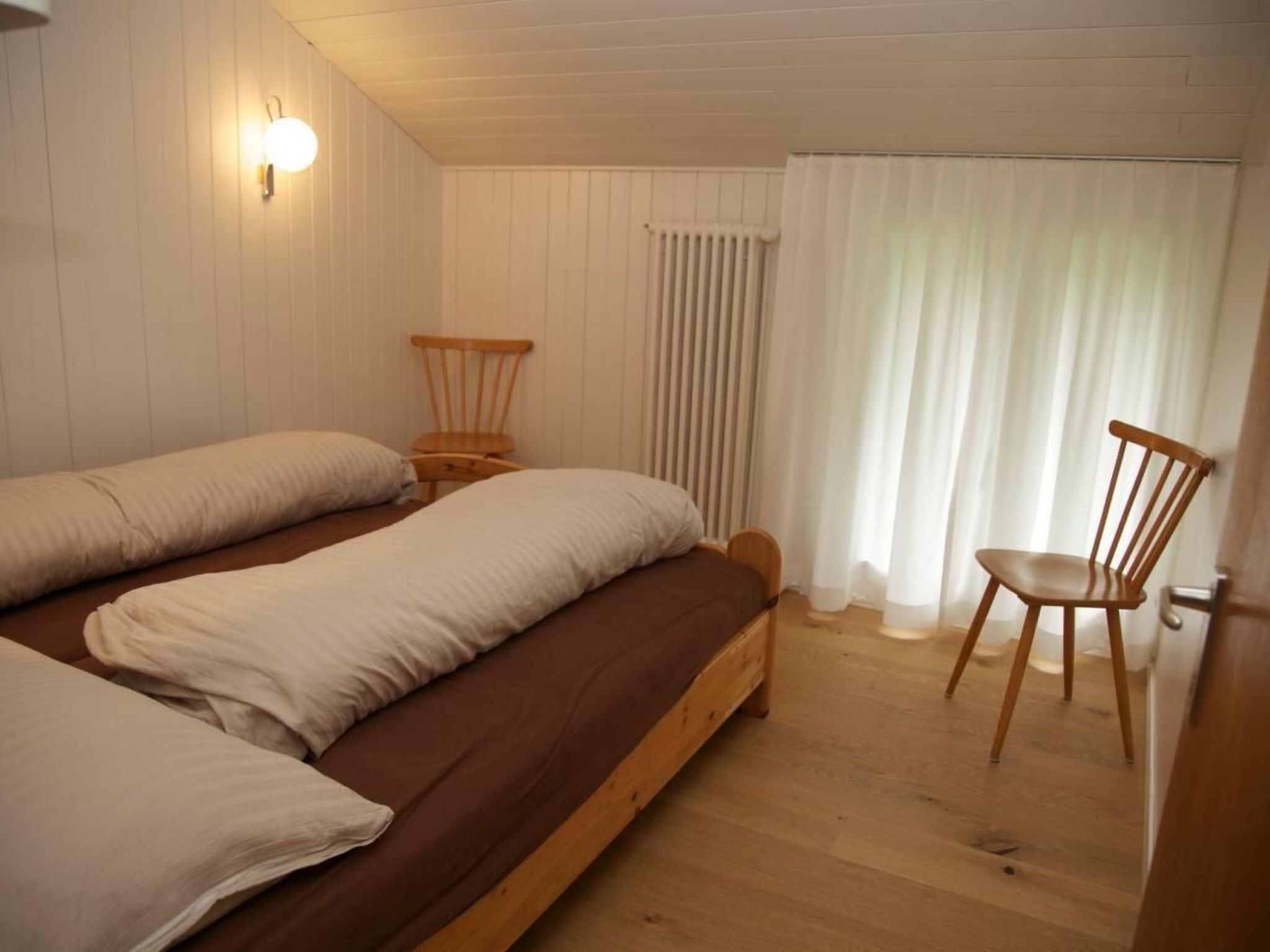 Una habitación con dos camas, una silla de madera, una mesita de noche, una lámpara de pared y una ventana con cortinas.