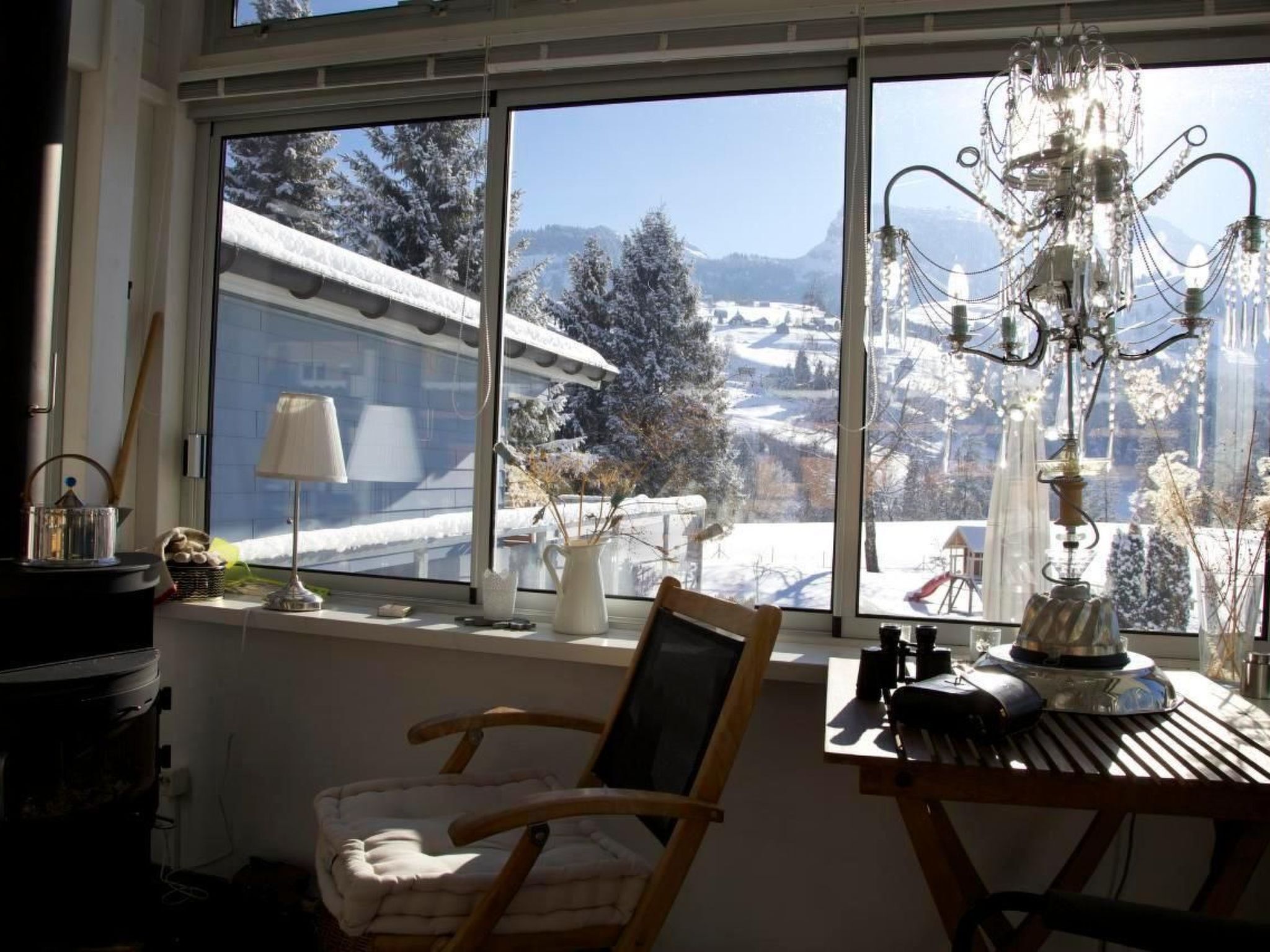 Een knus vertrek met een groot raam dat uitkijkt op een besneeuwd landschap, een houten stoel, een tafel met een lamp en een kroonluchter.