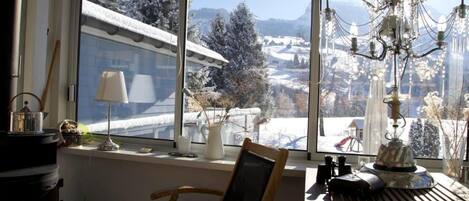 Een knus vertrek met een groot raam dat uitkijkt op een besneeuwd landschap, een houten stoel, een tafel met een lamp en een kroonluchter.