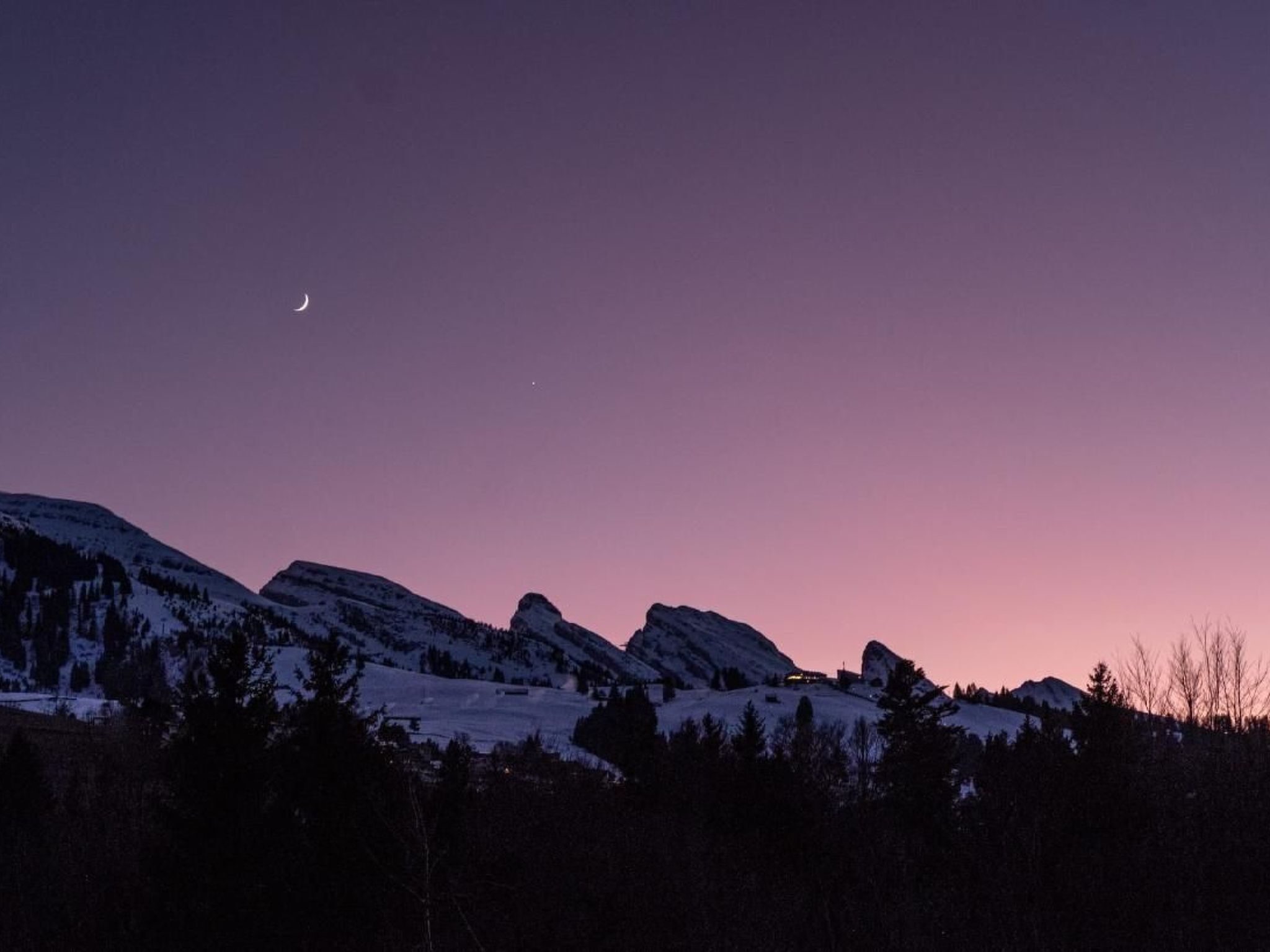 Un paysage montagneux au crépuscule, avec des sommets enneigés et un croissant de lune dans le ciel.