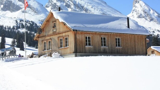 A wooden chalet with a snow-covered roof, set against a backdrop of snow-capped mountains. - Casa Uccello by Interhome (Wildhaus)