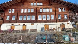 Un edificio de madera grande con múltiples ventanas y puertas, rodeado por una cerca y algunos materiales de construcción.