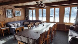 Una habitación acogedora con techo de madera, una gran mesa de comedor rodeada de sillas, un sofá y una ventana con vistas a árboles cubiertos de nieve.
