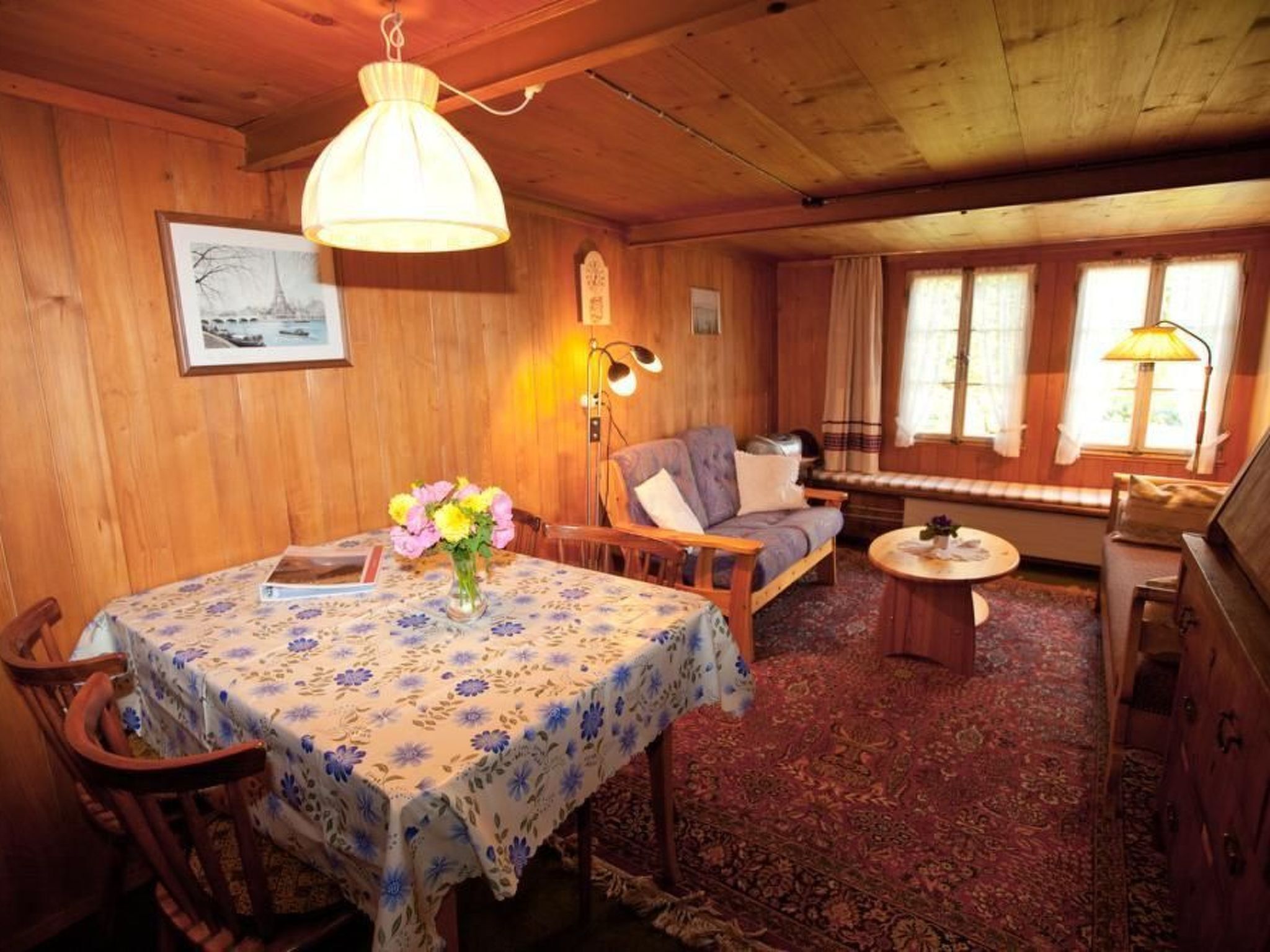 Une pièce chaleureuse avec un plafond en bois, une table à manger recouverte d’une nappe à motifs floraux, un canapé, une petite table ronde et un piano.
