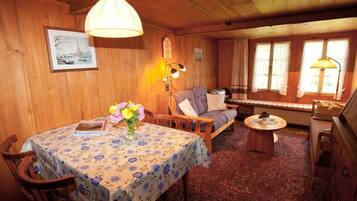 Une pièce chaleureuse avec un plafond en bois, une table à manger recouverte d’une nappe à motifs floraux, un canapé, une petite table ronde et un piano.