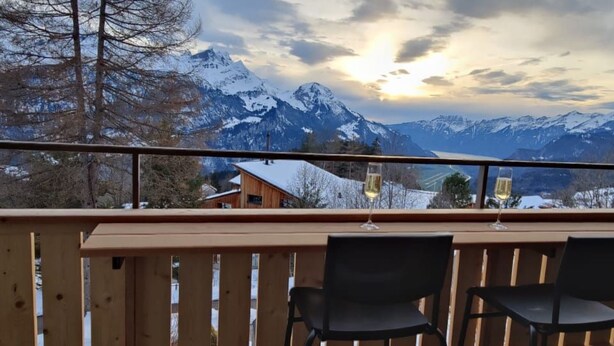 A balcony with wooden railings and two black chairs, overlooking a snowy mountain landscape with a building in the background. - Sunnehöckli by Interhome (Hasliberg)