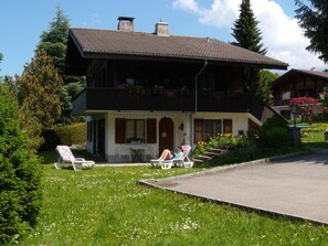 A two-story house with a balcony, a lawn with chairs, and a small table.