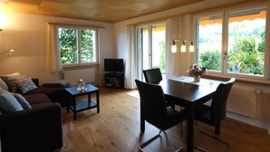 A living room with a sofa, TV, and dining table with chairs. There are large windows with curtains and a view of greenery outside. - Chalet Alouette by Interhome (Aeschi bei Spiez)