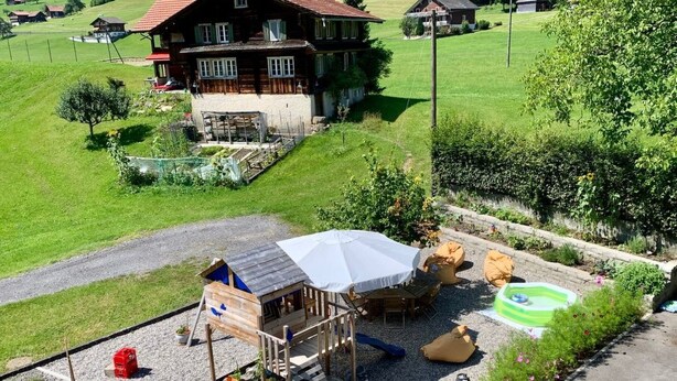 A wooden playhouse with a slide and a white umbrella, surrounded by a gravel area with potted plants and outdoor seating. - Gina's Apartment - Nähe zu See und Berge by Interhome (Sarnen)