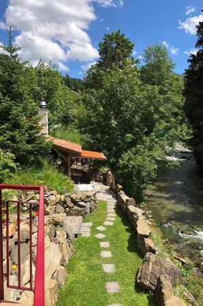 A stone pathway leading to a rustic wooden structure with a red roof, surrounded by lush greenery and a clear stream.