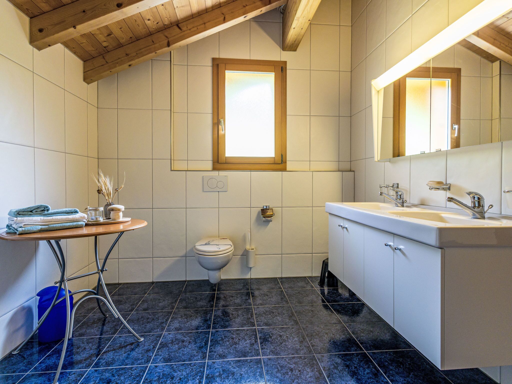 Une salle de bain avec des toilettes, un lavabo et un miroir, des poutres en bois au plafond et une fenêtre.