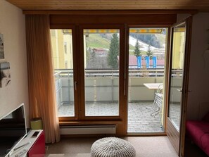 Una habitación con puerta de vidrio corredera que da a un balcón, televisión, una mesita y una cama roja.