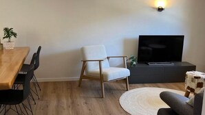 Un soggiorno moderno con un tavolo da pranzo in legno, una poltrona bianca, un divano grigio e una televisione a schermo piatto su un mobile scuro.