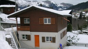 Un chalet in legno a due piani con un balcone, un tetto imbiancato dalla neve e uno sfondo montuoso.