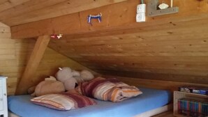 Een knus slaapkamer onder het dak met een houten schuine plafond, een enkel bed met een blauwe matras en gestreepte kussens, een witte dressoir en een kleurrijk vloerkleed.