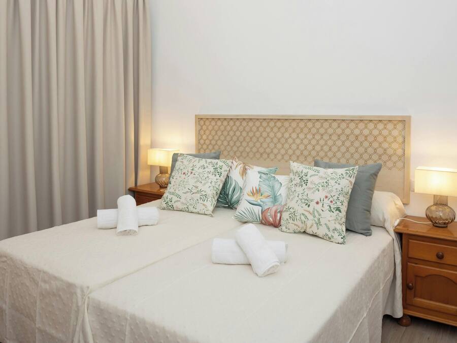 Una cama bien tendida con almohadas estampadas con motivos florales, dos lámparas de noche y una mesita de noche de madera con un cajón.
