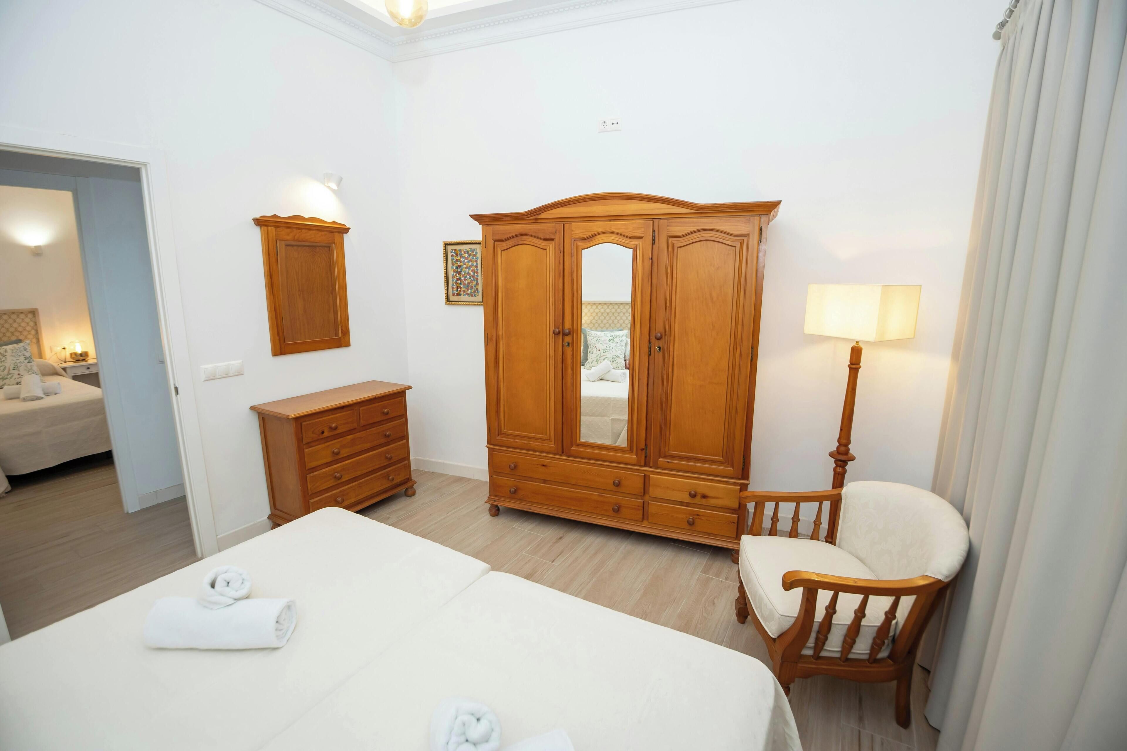 A hotel room with a large bed, a wooden wardrobe, a chair, a bedside table, and a lamp.