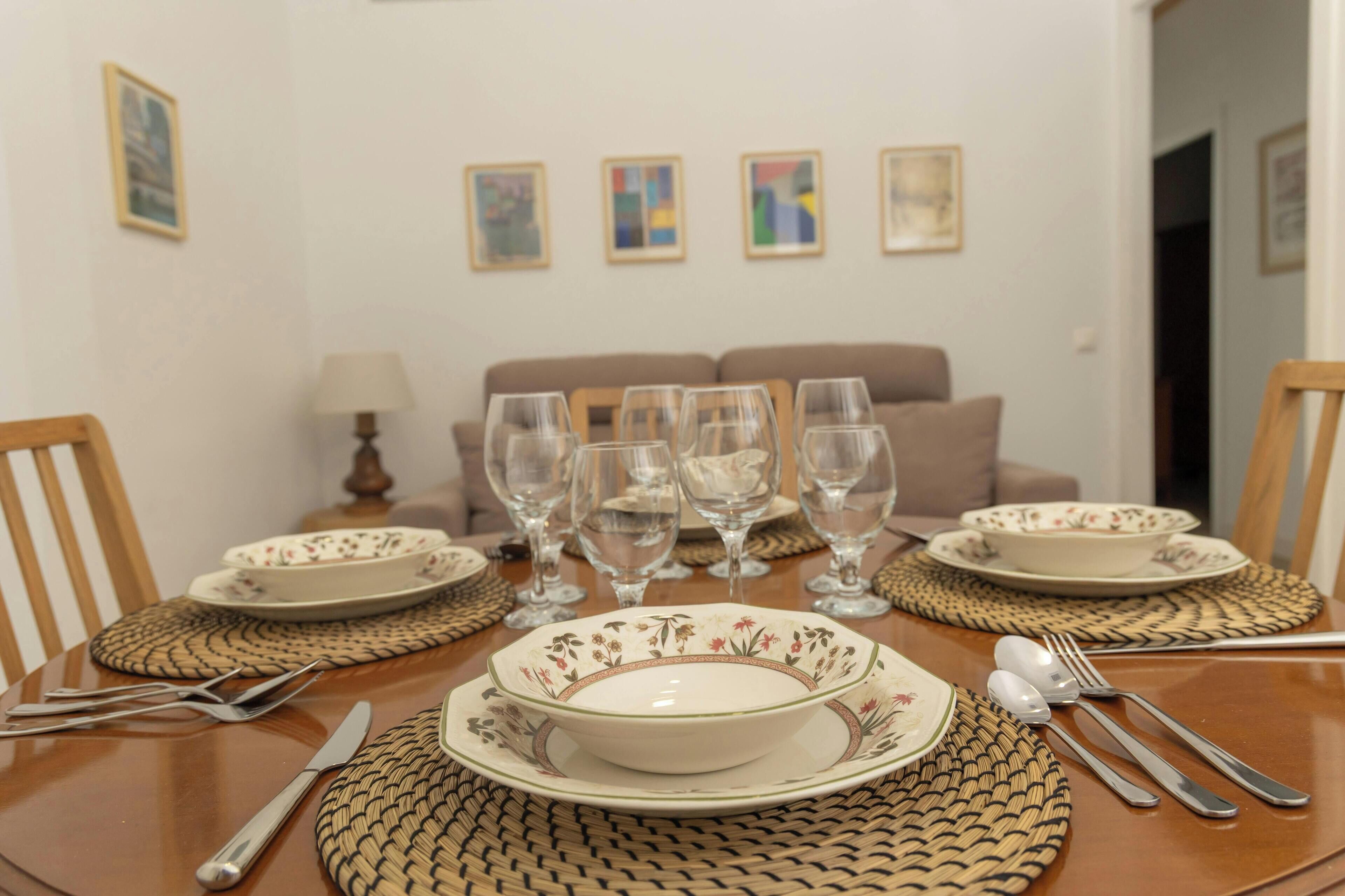 Una mesa de comedor dispuesta con platos, tazones, cubiertos y copas de vino, con un sofá y obras de arte en el fondo.