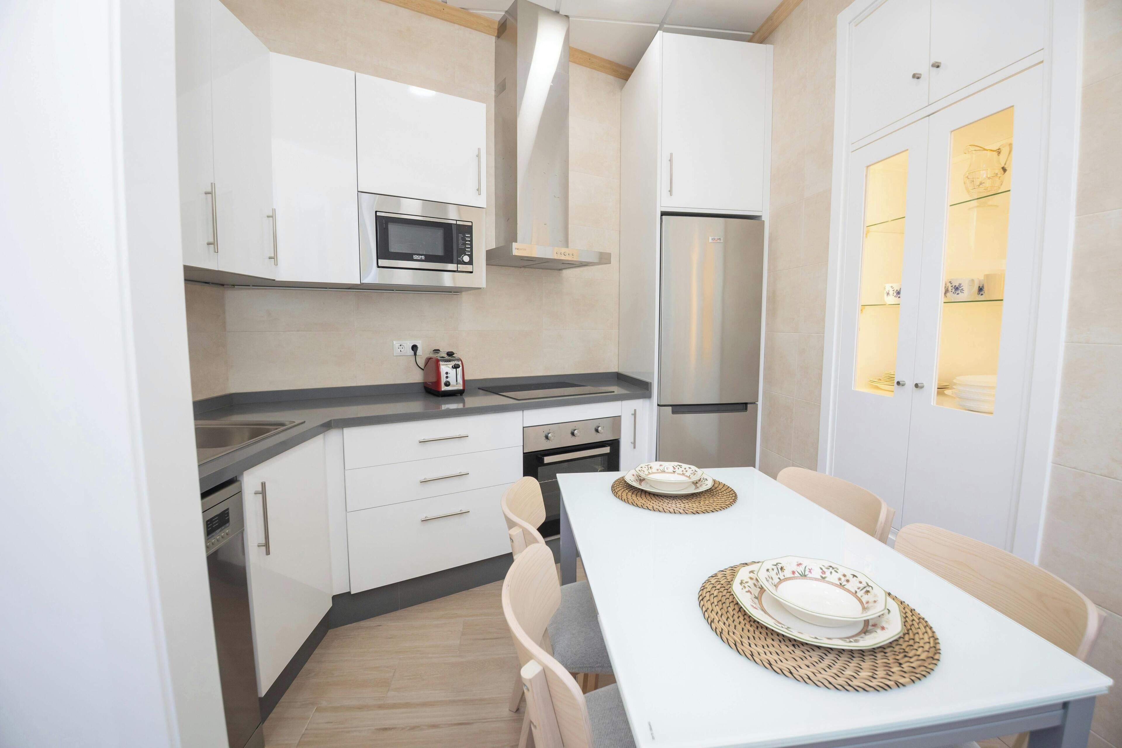 A compact kitchen with white cabinets, a microwave, a stove, and a refrigerator. There is a dining table with chairs and plates on it.