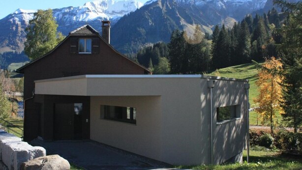 Una casa moderna de dos pisos con techo inclinado, chimenea y garaje. Está rodeada por un paisaje montañoso con picos nevados y bosques densos.