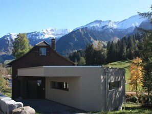 Una casa moderna de dos pisos con techo inclinado, chimenea y garaje. Está rodeada por un paisaje montañoso con picos nevados y bosques densos.