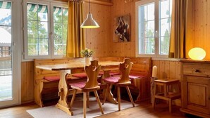 Een houten blokhut van binnen met een eettafel, stoelen, een hanglamp en grote ramen.