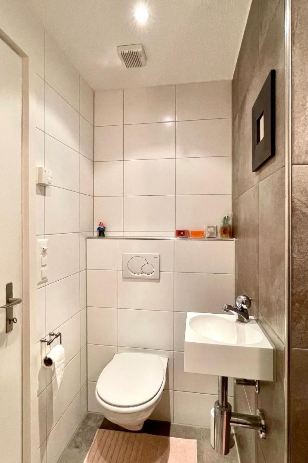 Un baño pequeño y moderno con azulejos blancos, un inodoro, un lavamanos y una ducha.